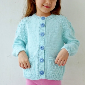以下が含まれることがあります： ボタン留めとポケットが2つ付いた薄い青色のニットカーディガン。カーディガンは質感のあるパターンで、柔らかな糸で作られています。