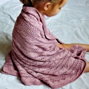 May include: A soft, pink knitted baby blanket with a cable knit pattern. The blanket is draped over a child sitting on a white bed.