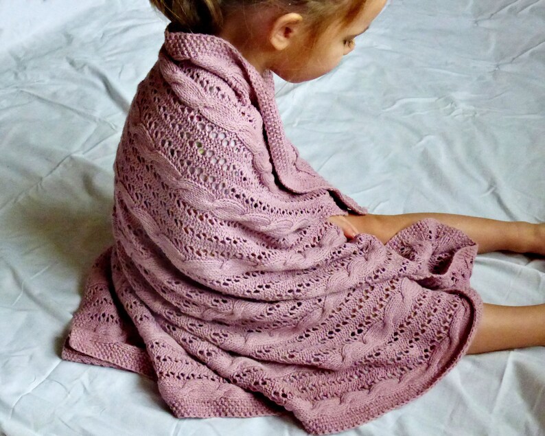May include: A soft, pink knitted baby blanket with a cable knit pattern. The blanket is draped over a child sitting on a white bed.
