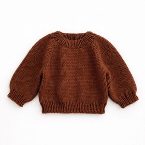 May include: A brown knit sweater with a crew neck and long sleeves. The sweater has ribbed cuffs and hem. The knitwear is a solid color and appears to be made of a soft yarn.