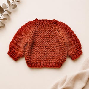 May include: A hand-knitted, crew-neck sweater in a gradient of orange and red hues. The sweater features a textured knit stitch and is likely made of a soft, warm yarn. The sleeves are set-in, and the overall style is classic and cozy.