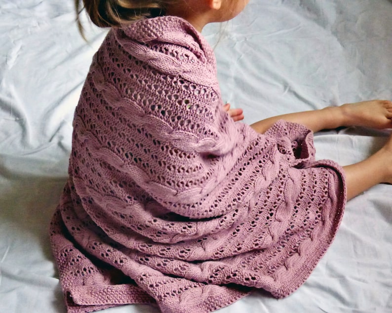 May include: A soft, pink knitted blanket with a cable knit pattern. The blanket is draped over a person sitting on a white surface.