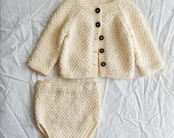 Könnte beinhalten: Cremefarbenes gestricktes Baby-Outfit, bestehend aus einer Knopf-Cardigan und passender Windelüberhose. Die Strickjacke hat lange Ärmel und braune Knöpfe. Das Set ist aus einem strukturierten Strickstoff gefertigt.