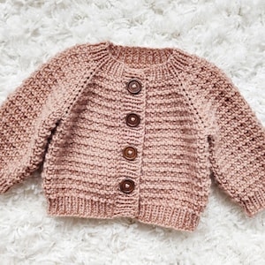 Peut inclure: Un cardigan en tricot rose pâle avec une fermeture boutonnée. Le cardigan a une texture côtelée et est fait avec un fil fin.