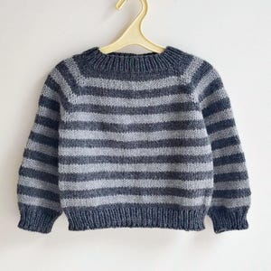 Puede incluir: Un suéter de punto de manga larga con rayas horizontales en tonos alternos de gris carbón oscuro y gris jaspeado claro. La prenda presenta un cuello redondo acanalado, puños acanalados y un dobladillo acanalado en el hilo gris carbón.