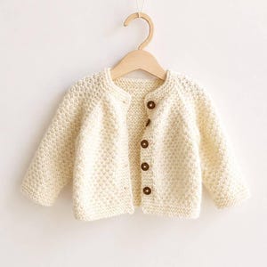 May include: Cream-colored knitted baby cardigan with long sleeves and a round neckline. The sweater features four brown buttons down the front and is displayed on a wooden hanger. The knitwear has a textured appearance.