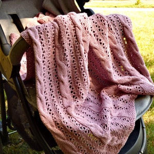 May include: A pink knitted baby blanket with a cable knit pattern, draped over a stroller.