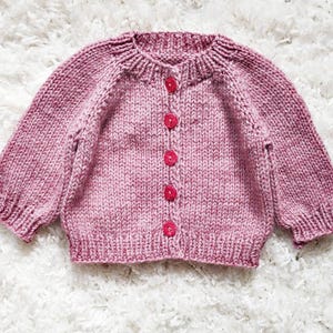 May include: A hand-knitted, dusty rose-colored cardigan sweater with long sleeves and a round neckline. The sweater features five bright pink buttons down the front. The sleeves have a slight puff at the shoulder and a ribbed cuff. The sweater is made of a soft, textured yarn.