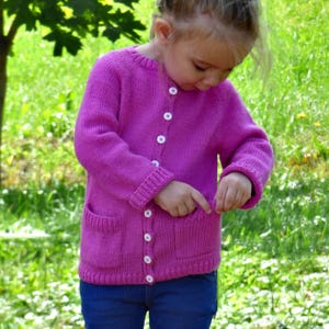 May include: A magenta knitted cardigan with two front pockets and white buttons. The long-sleeved sweater is worn by a child and paired with blue jeans. The background is a blurred green grassy area.