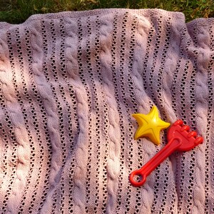 May include: A pink knitted blanket with a cable knit pattern. A yellow star and a red plastic rake are on top of the blanket.