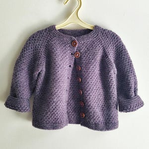 Peut inclure: Un gilet tricoté violet à manches longues avec un motif en maille texturée, une encolure ronde et une fermeture boutonnée sur le devant composée de six petits boutons ronds marron foncé. Le vêtement est présenté sur un cintre en plastique clair.
