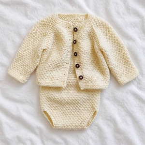 Puede incluir: Un conjunto de ropa de bebé de punto color crema, que incluye una rebeca con botones y un cubrepañales a juego. La rebeca tiene cinco botones marrones. El conjunto está hecho de tejido de punto texturizado y se muestra sobre una superficie blanca.