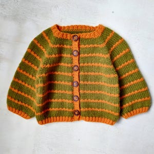 Può includere: Un cardigan in maglia a maniche lunghe con righe orizzontali verde oliva e arancione brillante. Il capo presenta uno scollo rotondo, polsini e orlo a coste e un'abbottonatura frontale verticale con sei bottoni rotondi marrone scuro.