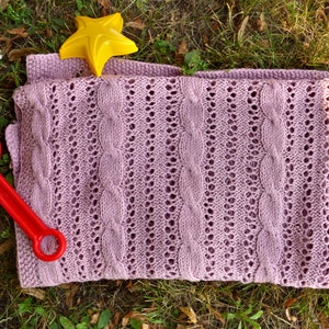 May include: A soft, pink knitted baby blanket with a cable knit pattern. The blanket is lying on a bed of green grass with a yellow star and a red plastic toy.