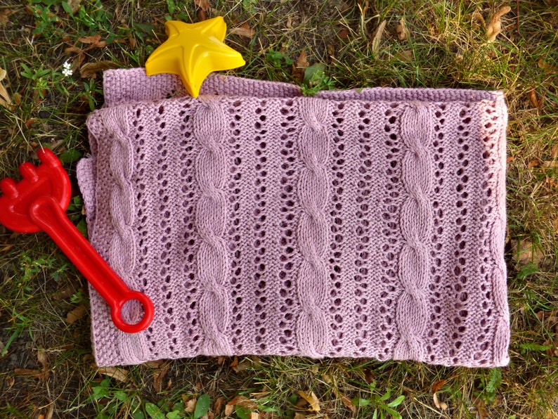 May include: A soft, pink knitted baby blanket with a cable knit pattern. The blanket is lying on a bed of green grass with a yellow star and a red plastic toy.