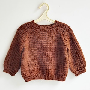 May include: A brown, hand-knitted sweater for a child, hanging on a white hanger. The sweater has a textured stitch pattern, a ribbed neckline, and ribbed cuffs. The sleeves are three-quarter length.