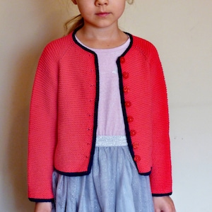 May include: A pink knitted cardigan with a black trim and red buttons. The cardigan has a slightly cropped length and long sleeves.