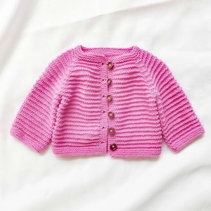 May include: A pink knitted baby cardigan with a ribbed design. The cardigan has long sleeves and is fastened with five brown buttons down the front. The garment is made of a soft, textured yarn and is displayed on a white background.