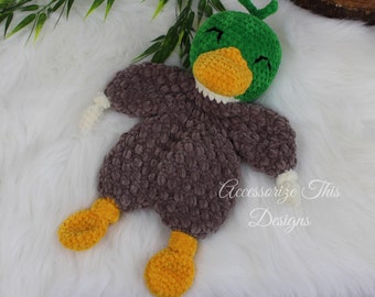 May include: A crocheted plush toy duck with a green head, brown body, white arms, and yellow feet. The duck has a smiling face with closed eyes.