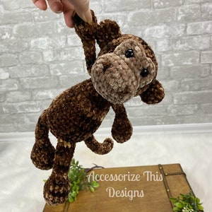May include: A brown crocheted monkey plush toy with black eyes. The monkey is hanging upside down with its arms and legs outstretched.