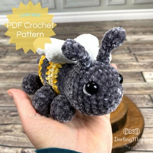 May include: A crocheted bee plush toy with gray and white fur, yellow and white striped body, and black eyes. The toy is being held in a hand. The text "LOW SEW PDF Crochet Pattern" is in a yellow starburst.