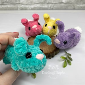 May include: Four crocheted plush bunny toys in different colors: teal, pink, yellow, and purple. The bunnies have black eyes and white noses. The teal bunny is being held in a hand.