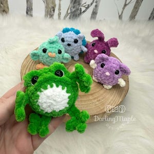 K&ouml;nnte beinhalten: Eine Sammlung von f&uuml;nf kleinen, geh&auml;kelten Pl&uuml;schtierfiguren, bestehend aus einem gr&uuml;nen Frosch, einem hellblauen Igel, einem violetten Hasen, einem hellvioletten Wesen und einem mintgr&uuml;nen Vogel, alle mit schwarzen Kunststoffaugen und weicher Textur.