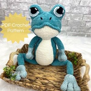 May include: PDF crochet pattern for a teal blue crocheted frog with white belly and large black eyes. The frog is sitting in a woven basket.