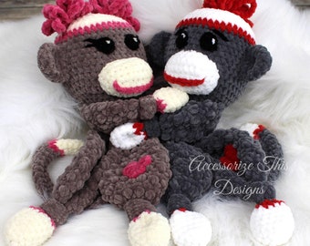 May include: Two crocheted sock monkey toys, one gray and one brown, with red and white accents. The monkeys are sitting on a white fluffy surface. The gray monkey has a red and white striped hat and the brown monkey has a pink and white striped hat.