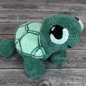 May include: A crocheted green turtle plush toy with a light green shell and large black eyes. The toy is sitting on a wooden surface.