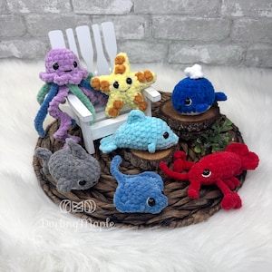 Puede incluir: Una colección de criaturas marinas hechas a mano con ganchillo, que incluye una medusa morada, una estrella de mar amarilla, una ballena azul, un tiburón gris, un delfín azul, un cangrejo rojo y una manta raya azul. Los juguetes están dispuestos sobre una estera tejida.