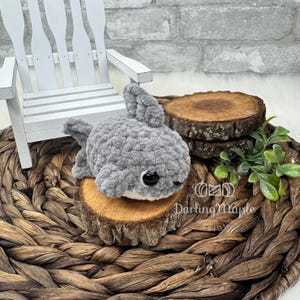 May include: A gray crocheted shark plush toy with black safety eyes and white belly. The shark is lying on a small wooden slice, set on a woven basket. A white wooden chair is in the background.