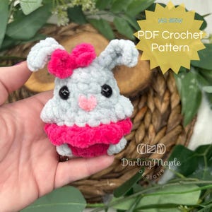 May include: A gray crocheted bunny with pink ears, a pink bow, and a pink skirt. The bunny is being held in a hand. The image includes the text "NO SEW PDF Crochet Pattern" and "Darling Maple Designs".