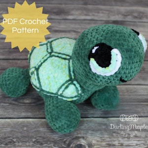 May include: A crocheted green turtle with a light green shell and large black eyes. The turtle is sitting on a wooden surface. The text "PDF Crochet Pattern" is in a yellow starburst.