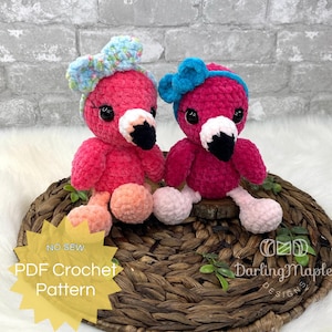 May include: Two pink crocheted flamingo toys with blue bows. The flamingo on the left has a floral print bow and the flamingo on the right has a solid blue bow. The toys are sitting on a woven basket with green leaves.