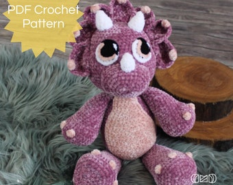 May include: PDF crochet pattern for a plush purple and pink triceratops dinosaur toy. The dinosaur has large eyes and a white horn. The pattern is available for download.