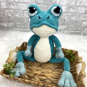 May include: A teal blue crocheted frog plush toy with white crocheted accents and large black eyes. The frog is sitting in a woven wicker basket.