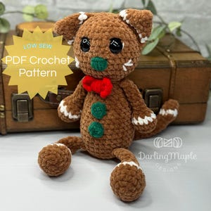 Può includere: Un gatto di pan di zenzero all'uncinetto marrone con un papillon rosso, bottoni verdi e dettagli bianchi. Il peluche ha occhi neri e una coda. Il testo sull'immagine recita "LOW SEW PDF Crochet Pattern".