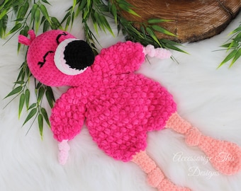 May include: A pink crocheted flamingo plush toy with black eyes, a white beak, and pink and orange legs. The toy is lying on a white fluffy surface.
