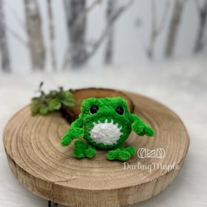 K&ouml;nnte beinhalten: Ein kleines, handgefertigtes H&auml;kel-Frosch-Stofftier aus leuchtend gr&uuml;nem Garn mit einem weichen wei&szlig;en Bauchfleck. Der Frosch hat zwei schwarze Kunststoff-Sicherheitsaugen und eine strukturierte, flauschige Optik. Der Text DarlingMaple DESIGNS ist sichtbar.