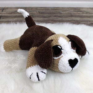 May include: A brown and white crocheted stuffed dog lying on a white fluffy surface. The dog has large black eyes and a black heart-shaped nose.