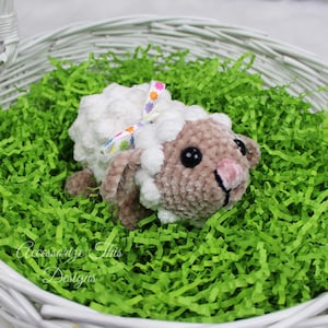 May include: A white and brown crocheted lamb with a colorful floral ribbon around its neck, sitting in a white wicker basket filled with green shredded paper.