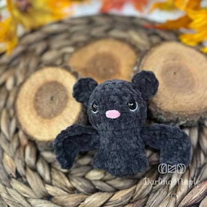 May include: A handmade, dark gray crocheted bat plush toy with a pink nose and embroidered eyes. The bat has outstretched wings and small ears. The plush is displayed on a woven basket and wooden slices. The text "Darling Maple Design" is visible.