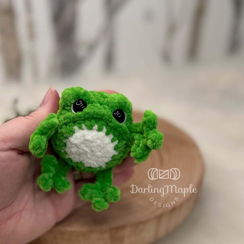 K&ouml;nnte beinhalten: Ein kleines, leuchtend gr&uuml;nes, geh&auml;keltes Frosch-Stofftier mit weicher, flauschiger Textur. Der Frosch hat einen runden wei&szlig;en Bauchfleck, zwei gro&szlig;e schwarze Kunststoff-Sicherheitsaugen mit wei&szlig;en Glanzlichtern und vier gespreizte Schwimmf&uuml;&szlig;e. Der Text DarlingMaple DESIGNS ist sichtbar.