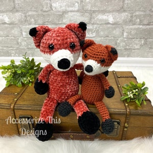 Peut inclure: Deux peluches de renard en crochet, l'une rouge et l'autre orange, assises sur une valise en bois. Le renard rouge est plus grand et a des yeux noirs et un nez blanc. Le renard orange est plus petit et a des yeux noirs et un nez blanc. La valise est brune et a un loquet en métal.