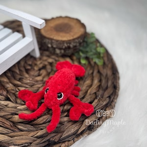 May include: A handmade red crochet lobster toy with black eyes and detailed claws. The plush toy is set on a woven brown surface, with a white wooden chair and a wood slice in the background. A whimsical design.