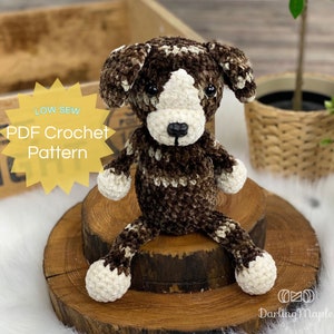 May include: A handmade crochet dog in brown and white, with a black nose and button eyes. The plush toy is sitting on a wooden slice. Text on the image reads "LOW SEW PDF Crochet Pattern".