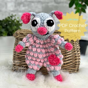 May include: A crocheted plush toy shaped like a creature with pink and grey fur. The toy has large black eyes, a pink nose, and pink ears. The toy is standing on a white fluffy surface. The text "LOW SEW PDF Crochet Pattern" is visible in the background.
