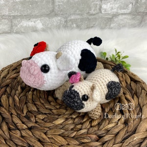 May include: Two crocheted stuffed cows, one white with black spots and a red flower, and one beige with black spots, sitting on a woven basket.