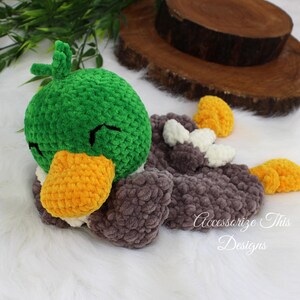 May include: A green, gray, and yellow crocheted duck plush toy with a closed eye expression. The duck is lying on its side with its head resting on its wing.